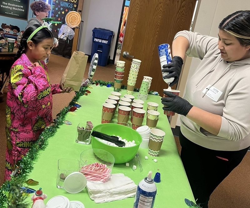 A female child in pink pajamas waits as a woman sprays whipped cream on top of a cup of hot chocolate.