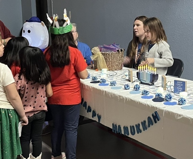 Children listen as a woman holding a small child speaks to them from behind a table featuring items associated with Hanukkah.