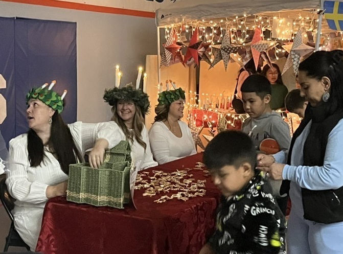 Children and adults file past a table featuring items associated with Christmas in Sweden. Three women seated behind the table wear white outfits and wreaths of candles on their heads.