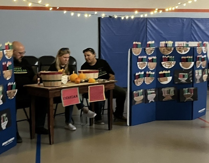 Three adults sit behind a table amidst a display detailing traditions associated with Kwanzaa.