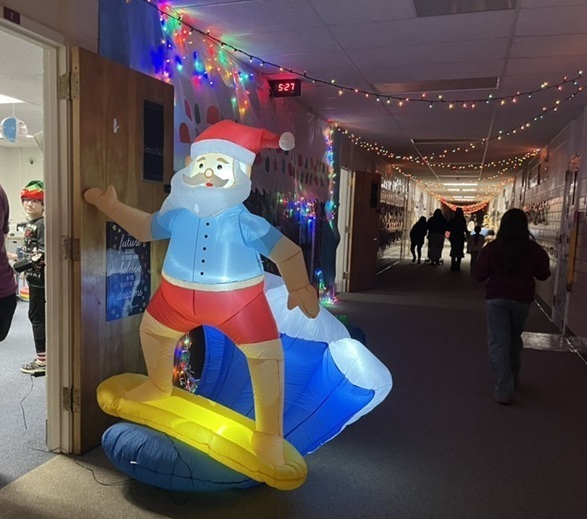 An inflatable Santa Claus riding a surfboard is seen outside a classroom along a Christmas-light-decorated hallway.
