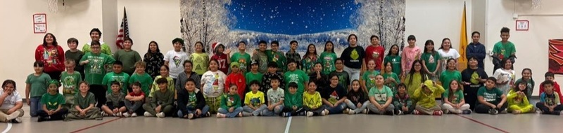 A large group of children wearing green and/or Grinch-related attire poses for a photo in a gym in front of a backdrop depicting a winter scene.