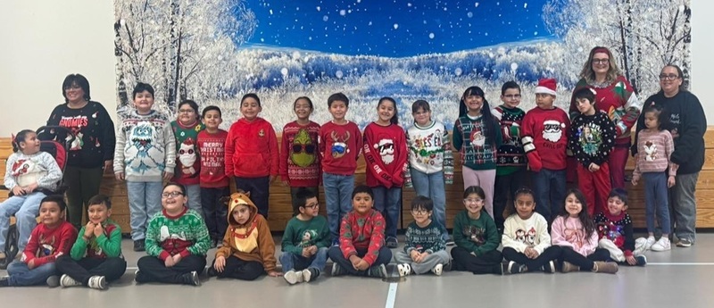 A large group of children wearing holiday-themed sweaters poses for a photo in front of a winter backdrop.