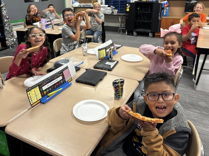 Children pose for a photo while eating pizza at their desks in a classroom.