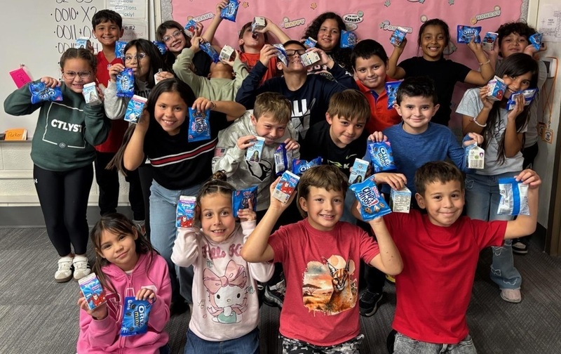 A group of children pose for a photo while holding up small bags of cookies and cartons of milk.