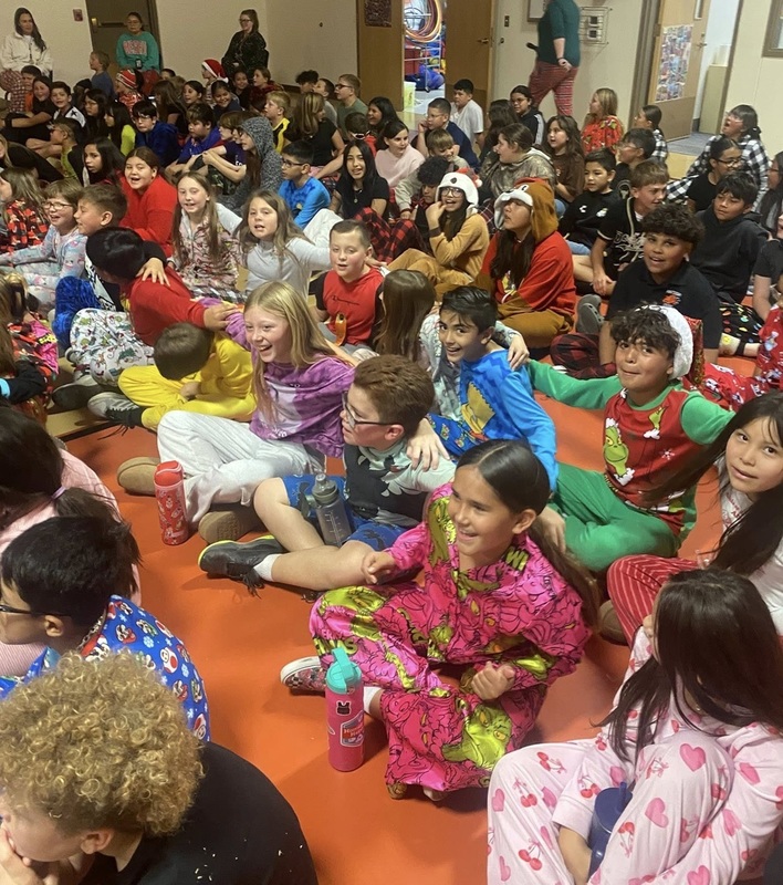 A large group of children in holiday pajamas sit on the floor of a gym.