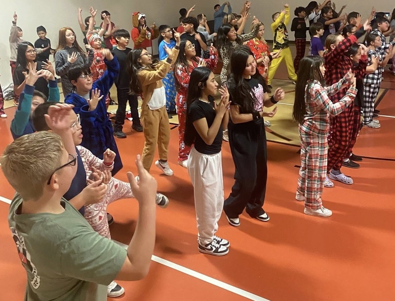 A large group of children wearing holiday pajamas stands in a gym and performs hand motions amidst a dance.