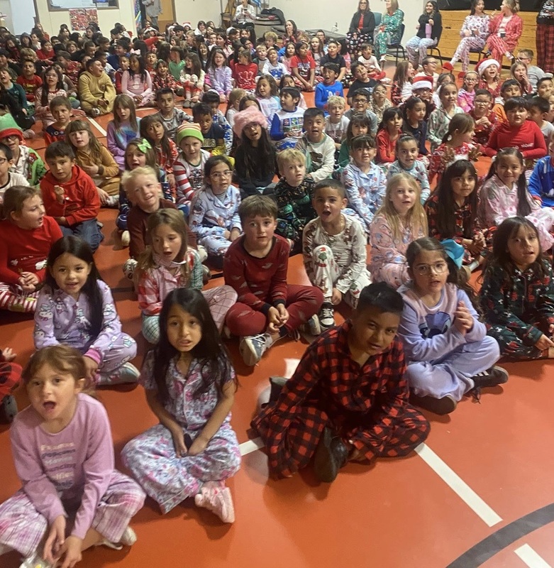 A very large group of children wearing holiday pajamas and seated on the floor of a gym look toward the camera as they sing a song.