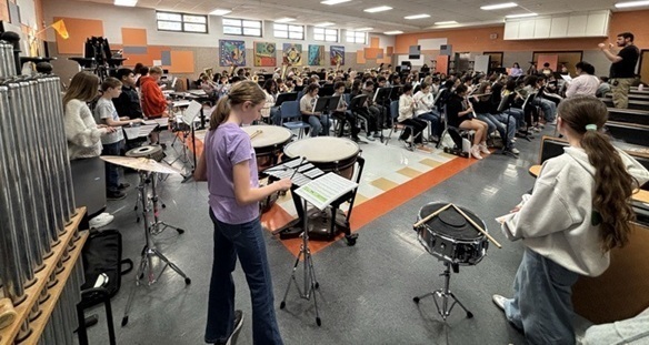 A large group of preteens is seen in a band hall, playing instruments as a male band director leads them.