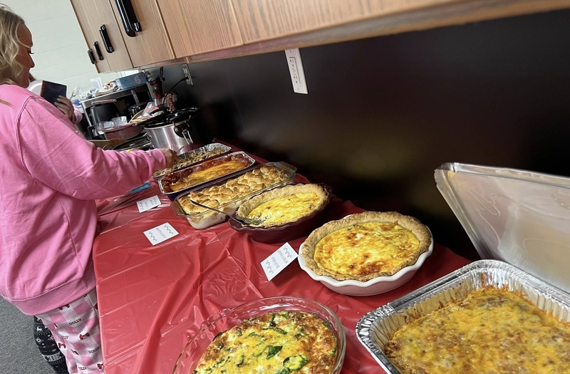 A female teacher in a pink top and pajama pants is seen walking along a buffet filled with quiches.