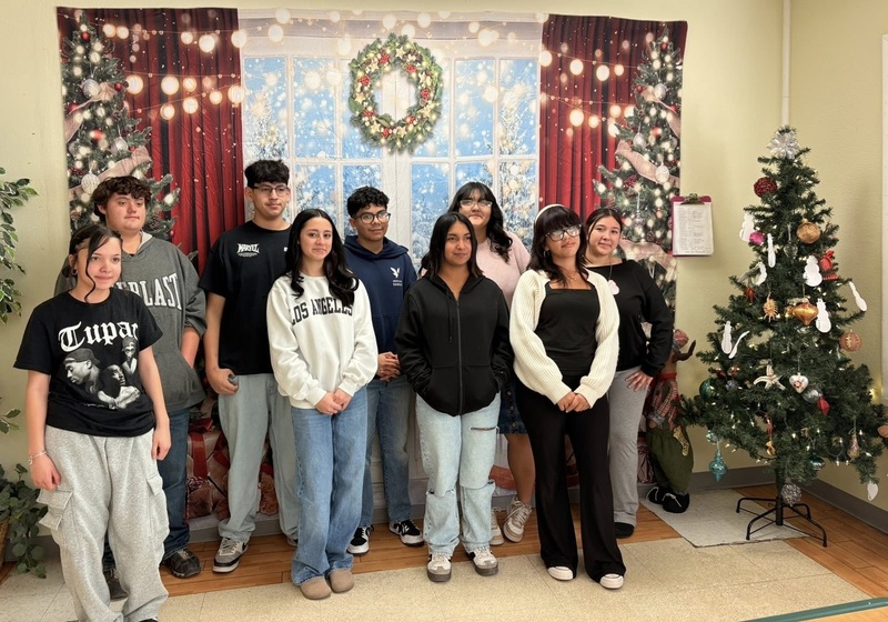 A small group of teens is seen standing in front of a winter window backdrop alongside a Christmas tree.