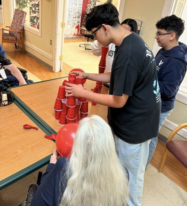 Two male teens are seen stacking red Solo cups for a game as two elderly residents of a nursing facility look on.