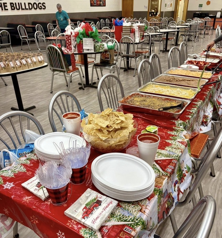 In a photo of a school cafeteria, one table is laden with enchiladas, chips and salsa, beans, rice, plates and napkins. Other tables in the distance hold goodie and gift bags.