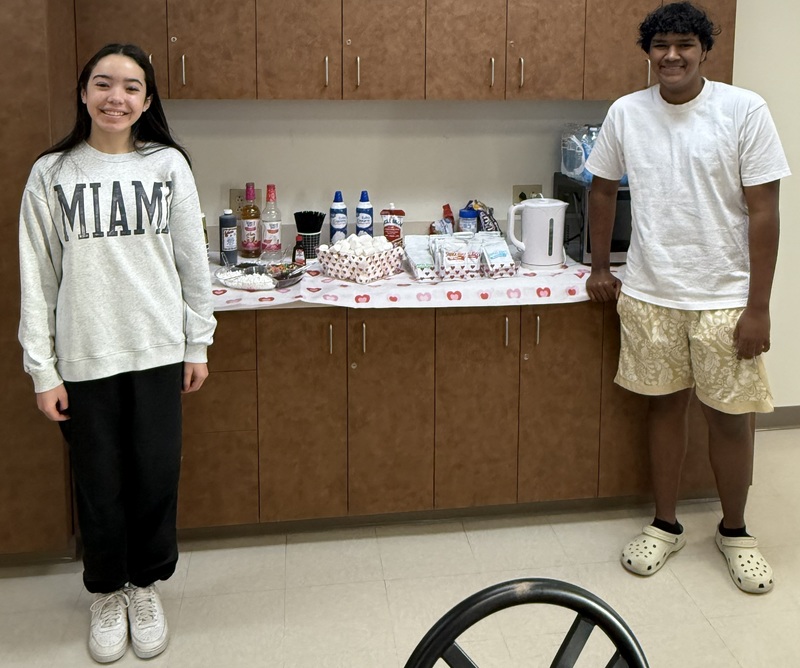 A female and male student smile for the camera while standing in front of a cabinet filled with hot chocolate supplies.