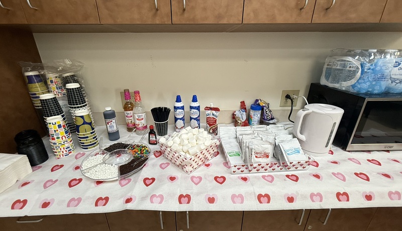A cabinet is seen laden with hot chocolate packets, marshmallows, candies, whipped cream, cups, and bottles of flavoring.
