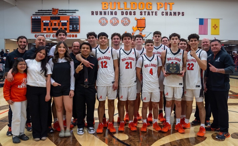 A boys' basketball team in white and orange uniforms poses for a picture alongside four male coaches, two female managers, and one female child. One of the players holds a trophy.
