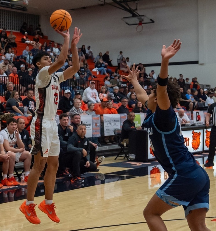 A teen boy in a white and orange basketball uniform is shown in mid-air as he releases the ball from his right hand. An opponent in blue has both arms in the air as he attempts to interfere.