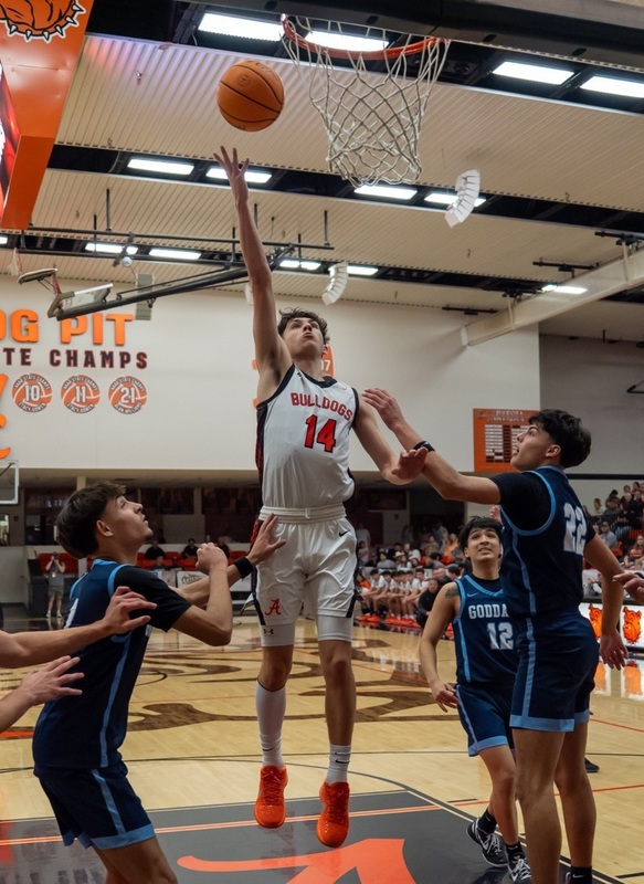 A teen boy in a white and orange basketball uniform jumps as he releases the balll from his right hand. Three opponents in blue watch from below.