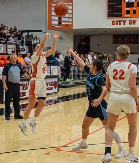 A teen girl in a white and orange basketball uniform is shown in mid-air as she releases the ball from both hands beyond the three-point arc. An opponent in blue stands in front of her with an arm raised.