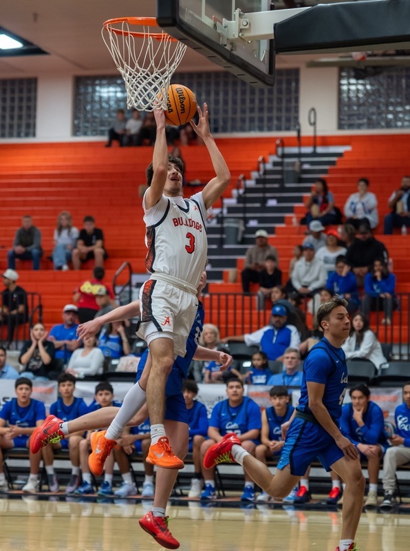 A teen boy in a white and orange basketball uniform is shown high off the court as he prepares to lay the ball off the glass. Two opponents in blue can be seen behind him.