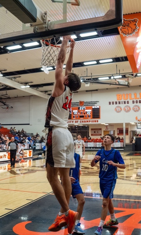 A teen boy in a white and orange basketball uniform is seen from the side as he dunks the ball. Two opponents in blue look on.