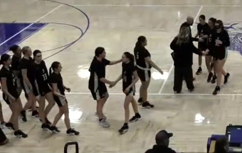 Two groups of teen girls in black and white basketball uniforms walk toward one another. Some shake hands and pat each other's backs.
