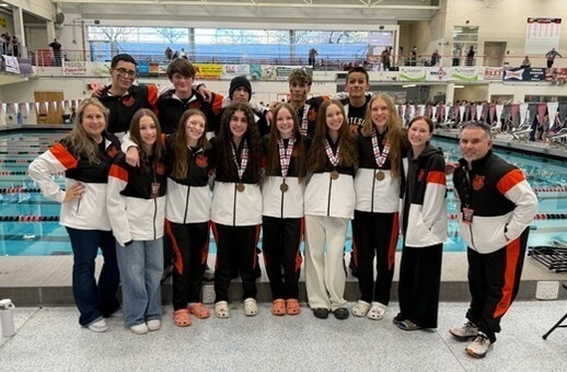 A group of teen girls and boys in orange, black and white swimming warm-ups, some wearing medals around their necks, pose for a photo with a female and male coach.