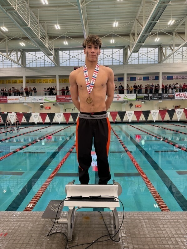 A teen boy in black and orange pants stands on a starting block in front of a pool wearing a bronze medal around his neck.