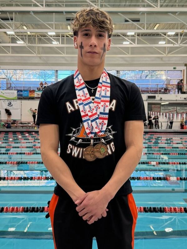 A teen boy in a black t-shirt reading "Artesia Swimming" and black and orange pants poses for a photo in front of a pool while wearing two bronze medals around his neck.