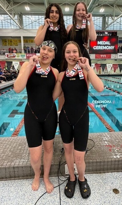 Four teen girls in black swimsuits pose for a photo while pretending to bite medals that are hanging around their necks.