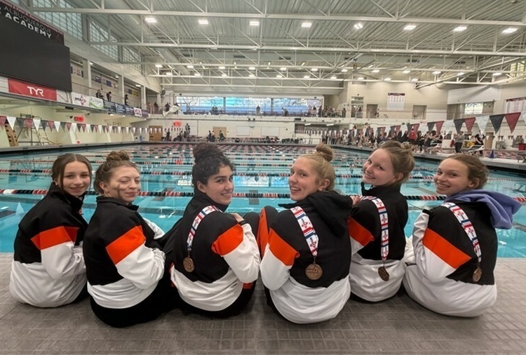 Six teen girls in orange, black and white swimming warm-ups and medals around their necks sit in front of a pool with their backs to the camera and turn their heads to look at the camera.