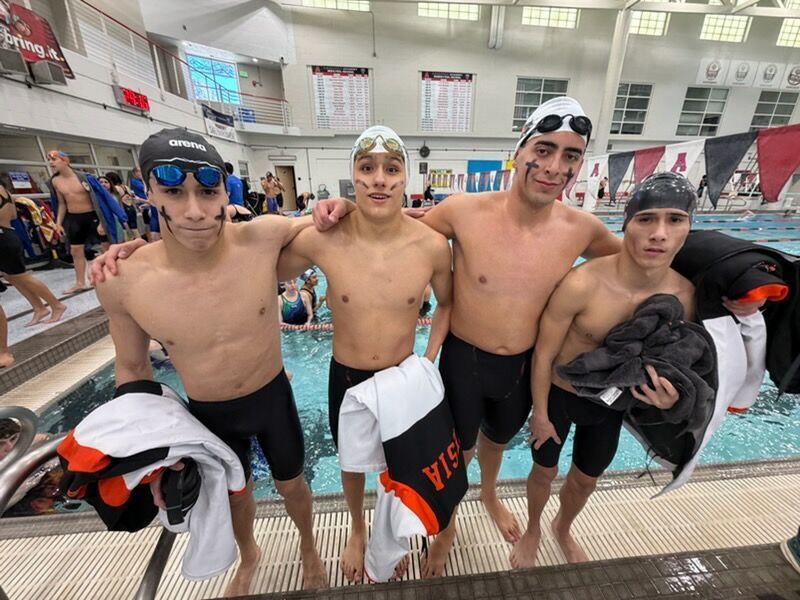 Four teen boys in black swim trunks, swim caps, and goggles pose for a photo alongside a pool.
