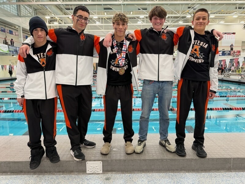 Five teen boys in orange, black and white swimming warm-ups pose for a photo alongside a pool with their arms over each other's shoulders.