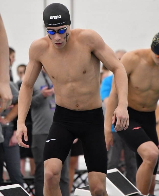 A teen boy in black swim trunks, a black swim cap, and goggles leans forward slightly as he climbs onto a starting block.