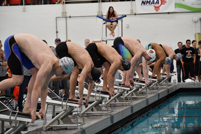 A row of teen boys in swim trunks bend down to grasp the front of their starting blocks as they prepare to dive into the pool.