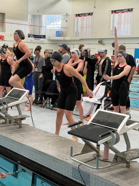 Four teen girls in black swimsuits stand behind starting blocks and move their arms as they prepare for the start of a race.