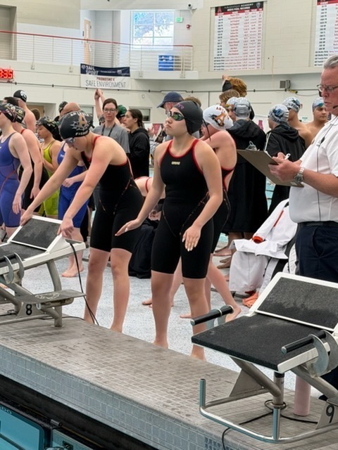 Four girls in black swimsuits, black swim caps, and goggles stand behind starting blocks and move about as they prepare for the start of a race.