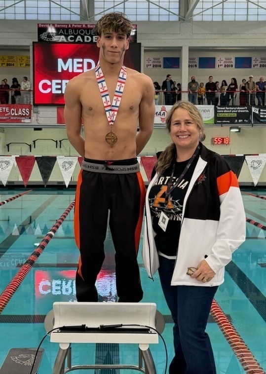 A teen boy in black and orange pants with a bronze medal around his neck poses atop a starting block. A female coach in an orange, black and white jacket smiles as she stands beside him on the ground.