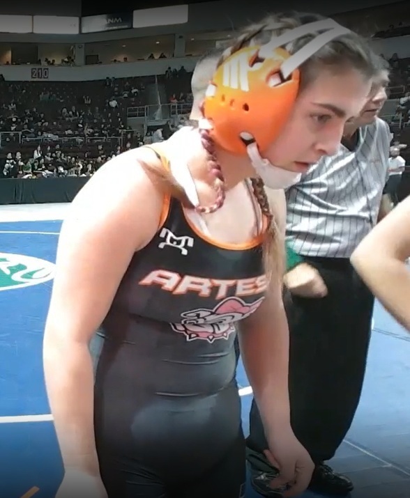 A teen girl in a black wrestling uniform and orange ear guards stands and listens to someone out of view speak.