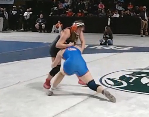 A teen girl in a black wrestling uniform and orange ear guards leans over the back of an opponent in blue who is trying to take out her legs.