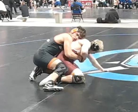 A teen boy in a black wrestling uniform and orange ear guards hangs onto the back of an opponent in maroon who is struggling to escape.