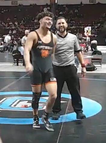 A teen boy in a black wrestling uniform laughs with a referee as he walks off the mat.