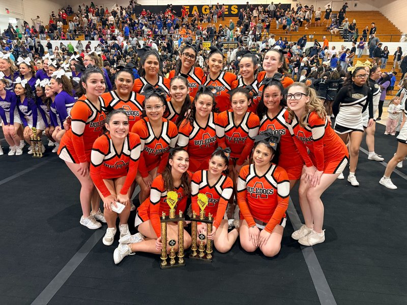 A large group of teen girls in orange and white cheerleading uniforms poses for a photo on a large black mat with other cheerleaders and a crowd behind them. In front of them sit two trophies.
