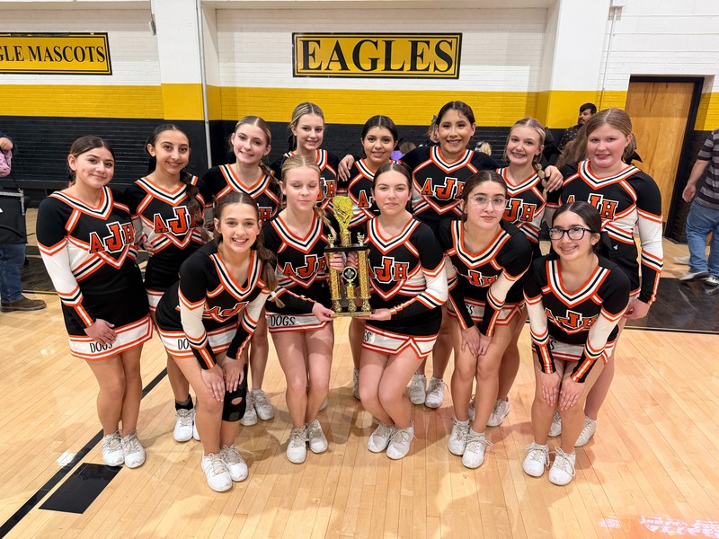 A group of teen girls in black, orange and white cheerleading uniforms poses for a photo. Two girls in the center hold a trophy.