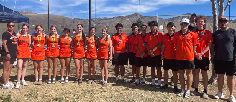 Seven female and seven male teens, all in orange tennis uniforms, pose for a photo with a female coach and two male coaches. Some of the teens wear medals around their necks. Mountains can be seen in the background.