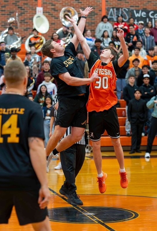 A male coach in black leaps high in the air above a teen basketball player in orange and black during the opening tip-off.