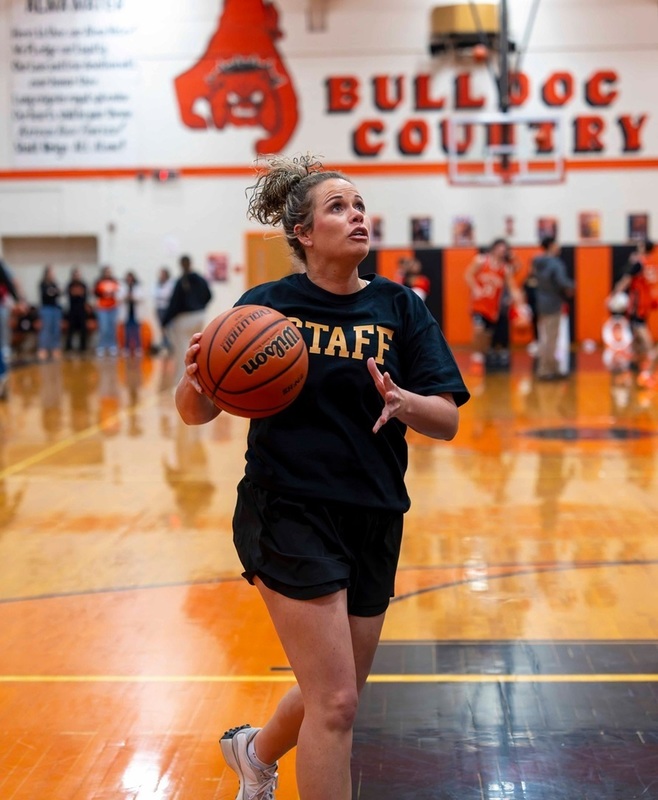 A woman in black shorts and a black shirt reading "Staff" in orange shifts the basketball to her right hand as she looks up at the basket.