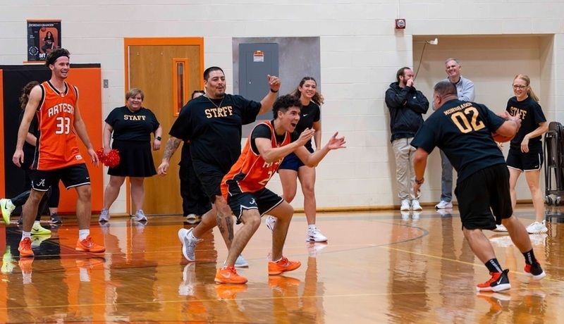 A male coach in black shorts and a black shirt with an orange "02" on he back prepares to dribble around a teen boy in orange and black as another teen and another male coach look on and smile.
