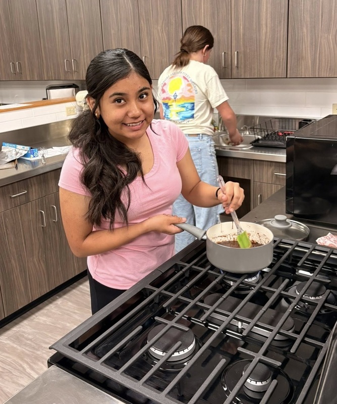 A teen girl smiles for the camera as she stirs a pot containing a chocolate liquid on a gas stovetop.