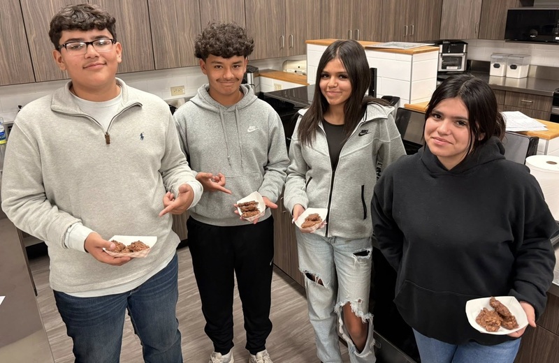 Two teen boys and two teen girls pose for a photo in a kitchen classroom. The students hold small paper trays containing chocolate cookies.